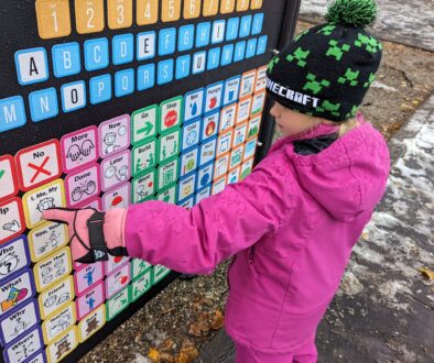 A student wearing a pink jacket and snow pants points to a square on a communication board