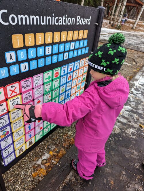 A student wearing a pink jacket and snow pants points to a square on a communication board