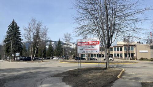 In the foreground of the photo is a sign with a white background and blue and red writing that reads: Queen Victoria Hospital and Health Centre emergency drop off. In the background of the photo is a parking lot with cars parked in front of a beige coloured building. There are also trees and a mountain covered in snow.